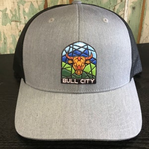 May include: A gray and black trucker hat with a black mesh back. The front of the hat features a colorful bull head design with a stained glass effect and the text "BULL CITY".