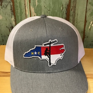 May include: A gray and white trucker hat with a patch of the North Carolina state flag with a silhouette of a lineman climbing a utility pole. The patch says "N*C" in yellow letters.