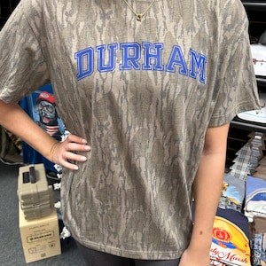 Op de afbeelding: Een T-shirt met korte mouwen met een camouflagepatroon in bruin- en grijstinten. Het woord "DURHAM" staat in blauwe blokletters op de borst. Het shirt is gemaakt van een zacht materiaal.