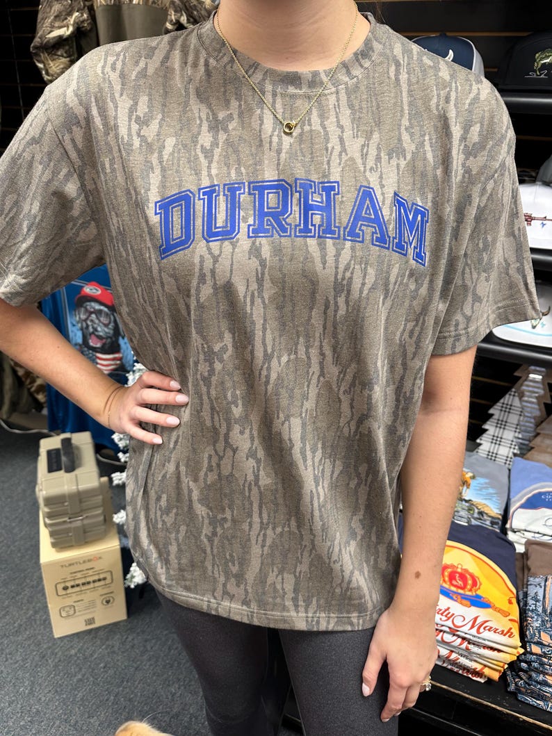 Op de afbeelding: Een T-shirt met korte mouwen met een camouflagepatroon in bruin- en grijstinten. Het woord "DURHAM" staat in blauwe blokletters op de borst. Het shirt is gemaakt van een zacht materiaal.