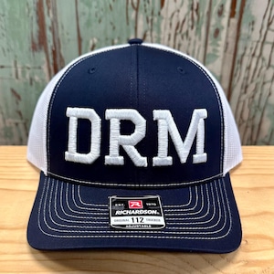 May include: A navy blue and white trucker hat with a white embroidered "DRM" on the front. The hat has a black adjustable strap with a white "Richardson 112" label.