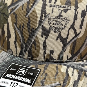 May include: Camouflage pattern trucker hat with the text "Treesland" and "Mossy Oak" logo. The hat has a Richardson tag that reads "Original 112 Trucker Adjustable". The hat is made of a durable fabric.