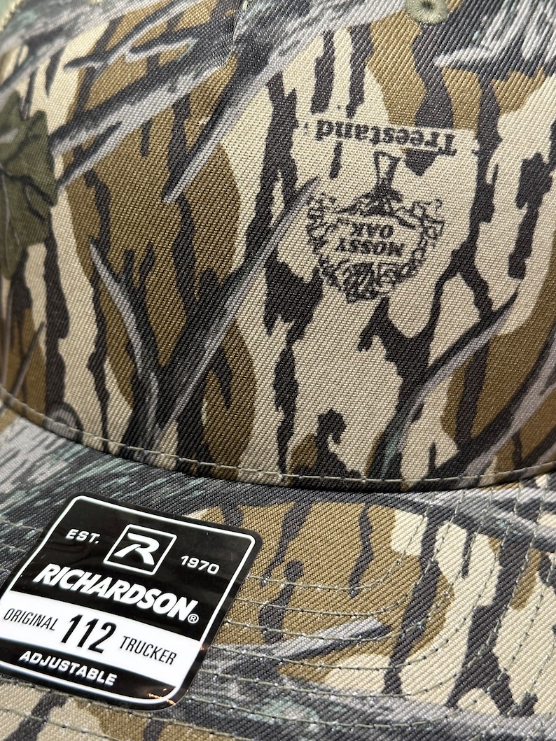 May include: Camouflage pattern trucker hat with the text "Treesland" and "Mossy Oak" logo. The hat has a Richardson tag that reads "Original 112 Trucker Adjustable". The hat is made of a durable fabric.