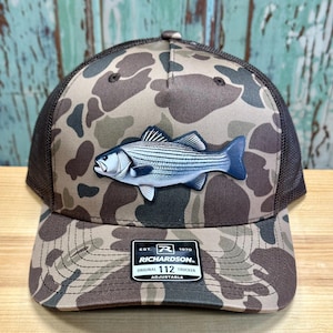May include: A brown and black camouflage trucker hat with a white fish patch. The hat has a black mesh back and an adjustable strap. The hat has the text "Richardson. Original Trucker 112 Adjustable" on the front.