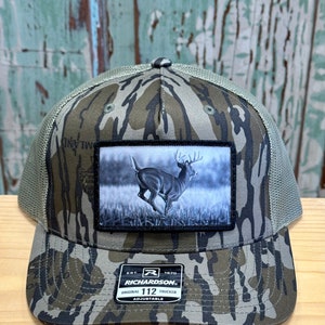 May include: A camouflage trucker hat with a black patch featuring a whitetail deer in a field. The hat has a black label with the text "Richardson" and "Original 112 Trucker Adjustable".