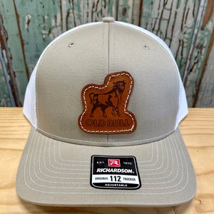 May include: A tan and white trucker hat with a brown leather patch featuring a bull and the text "Old Bull". The hat has a white mesh back and a black label with the text "Est. 1970 Richardson. Original 112 Trucker Adjustable".