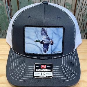 May include: A grey and white trucker hat with a patch featuring a mallard duck in flight. The patch has the text "AG Outfitters". The hat has a black label with the text "EST. 1970 RICHARDSON. ORIGINAL 112 TRUCKER ADJUSTABLE".