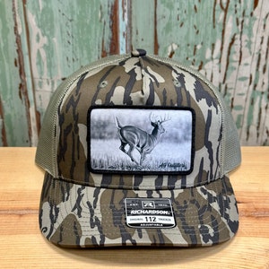 May include: A camouflage trucker hat with a black and white patch of a running deer. The patch has the text "AG Outfitters". The hat has the text "Richardson's Original 112 Trucker Adjustable" on the front.