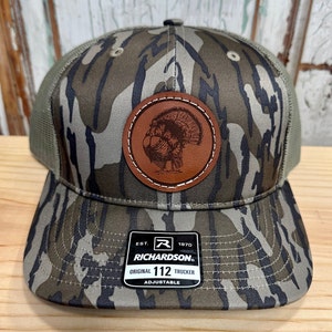 May include: Camouflage trucker hat with a brown leather patch featuring a turkey illustration. The hat has a mesh back and a curved brim. The tag reads "RICHARDSON ORIGINAL 112 TRUCKER ADJUSTABLE."