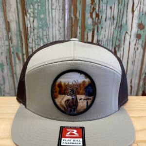 May include: A tan and brown flat bill snap back hat with a circular patch featuring a white-tailed deer in a field with a cabin in the background. The patch has the text "AC Designs" on the bottom.