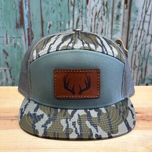 May include: A green and brown camouflage trucker hat with a brown leather patch featuring a deer antler design.