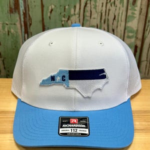 May include: White and light blue trucker hat with a North Carolina state outline and the letters "N.C." in blue. The hat has a mesh back and an adjustable strap with the Richardson logo and "112 Trucker" text.