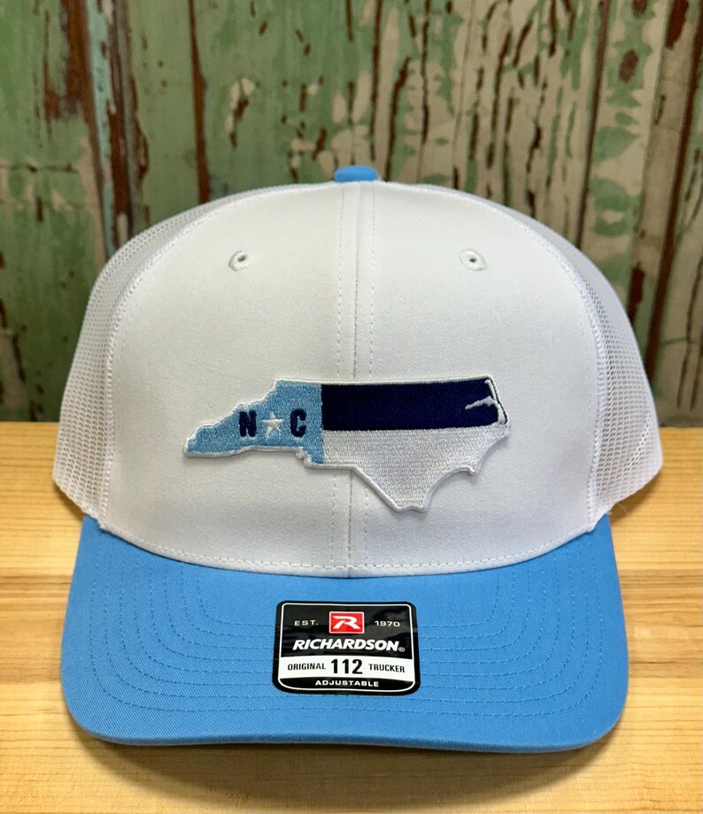 May include: White and light blue trucker hat with a North Carolina state outline and the letters "N.C." in blue. The hat has a mesh back and an adjustable strap with the Richardson logo and "112 Trucker" text.