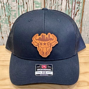 May include: Black trucker hat with a brown leather patch featuring a bull's head with a skyline and the letters "N.C." inside. The hat has a black mesh back and a red, white, and black label that reads "Richardson. Est. 1970 Original 112 Trucker Adjustable."