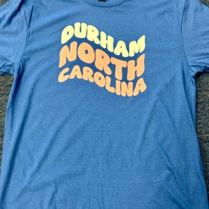 May include: A blue t-shirt featuring the text "Durham North Carolina" in a retro, wavy design. The text is in yellow and orange. The shirt is made of a soft material.