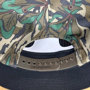 K&ouml;nnte beinhalten: Eine Camouflage-Baseballkappe mit schwarzem Schirm und Mesh-R&uuml;ckseite. Die Kappe hat einen braunen, verstellbaren Riemen mit dem eingepr&auml;gten Markennamen "RICHARDSON". Das Camouflagemuster umfasst Gr&uuml;n-, Braun- und Beiget&ouml;ne.