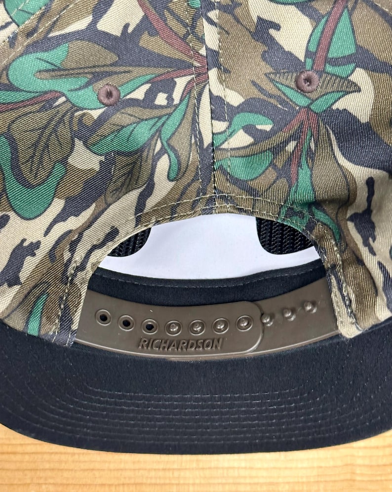 K&ouml;nnte beinhalten: Eine Camouflage-Baseballkappe mit schwarzem Schirm und Mesh-R&uuml;ckseite. Die Kappe hat einen braunen, verstellbaren Riemen mit dem eingepr&auml;gten Markennamen "RICHARDSON". Das Camouflagemuster umfasst Gr&uuml;n-, Braun- und Beiget&ouml;ne.