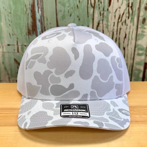 May include: A white and gray camouflage pattern trucker hat with a mesh back. The hat features a curved brim and an adjustable strap. The label reads "RICHARDSON 112 TRUCKER".