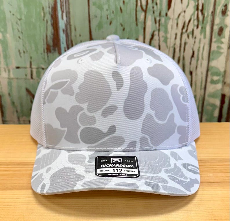 May include: A white and gray camouflage pattern trucker hat with a mesh back. The hat features a curved brim and an adjustable strap. The label reads "RICHARDSON 112 TRUCKER".