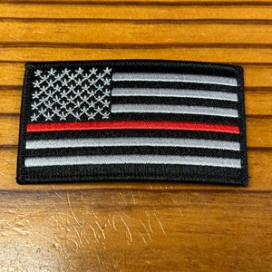 May include: A black and white American flag patch with a single red stripe across the middle. The patch is made of embroidered fabric.