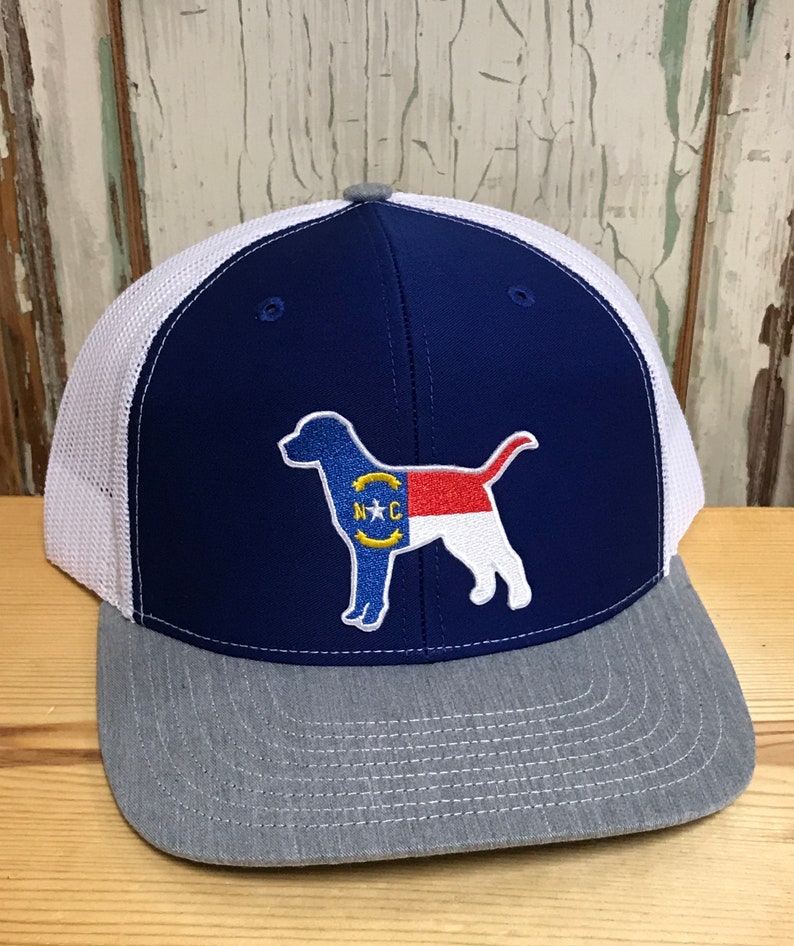 May include: A blue and white trucker hat with a grey brim. The hat features an embroidered patch of a dog silhouette with the North Carolina state flag design. The patch says "N.C."