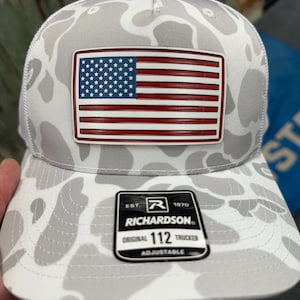Puede incluir: Gorra de camionero con un patrón de camuflaje blanco y gris. La gorra presenta un parche rectangular con la bandera estadounidense y la etiqueta de la marca Richardson. La gorra es ajustable.