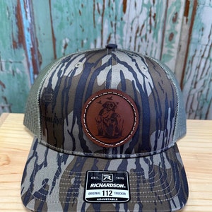 May include: A camouflage trucker hat with a brown leather patch featuring a dog and birds. The patch has white stitching around the edge. The hat has a black mesh back and a brown adjustable strap with the text "Richardson 112 Original Trucker Adjustable" and a logo.