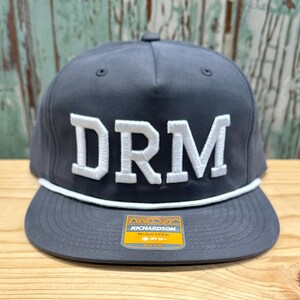 May include: A gray and white baseball cap with the letters "DRM" embroidered in white on the front. The cap has a white stripe around the brim and a yellow label with the text "Richardson Active UPF 50+ One Size" on the side.