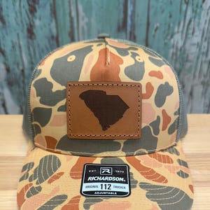 May include: A camouflage trucker hat with a leather patch featuring the outline of South Carolina. The hat has a mesh back and a curved brim. The front of the hat has a pattern of orange, gray, and tan shapes. The hat has the text "RICHARDSON" and "ORIGINAL 112 TRUCKER".