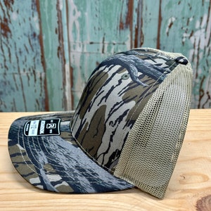 May include: A camouflage baseball cap with a curved peak and a mesh back. The cap features a pattern of green, brown, and grey, with a beige mesh back. The peak has a small tag with the text "RICHARDSON 112".