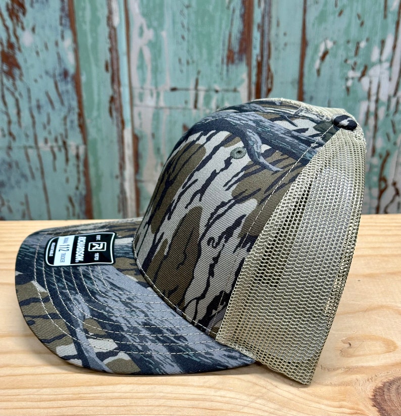 May include: A camouflage baseball cap with a curved peak and a mesh back. The cap features a pattern of green, brown, and grey, with a beige mesh back. The peak has a small tag with the text "RICHARDSON 112".