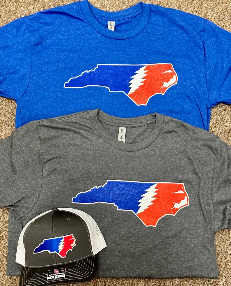 May include: Two t-shirts with a graphic of the state of North Carolina with a Grateful Dead lightning bolt design. One shirt is blue and the other is gray. A black and white baseball cap with the same graphic is also shown.