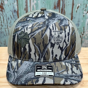 May include: A camouflage trucker hat with a curved peak and mesh back. The hat features a Mossy Oak Treestand pattern and a Richardson patch. The hat is adjustable and has a button on top.