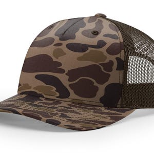 May include: A brown and tan camouflage trucker hat with a curved brim. The front panels feature a duck camouflage pattern, while the back panels are a dark brown mesh. The hat has a standard six-panel design and a button on top.