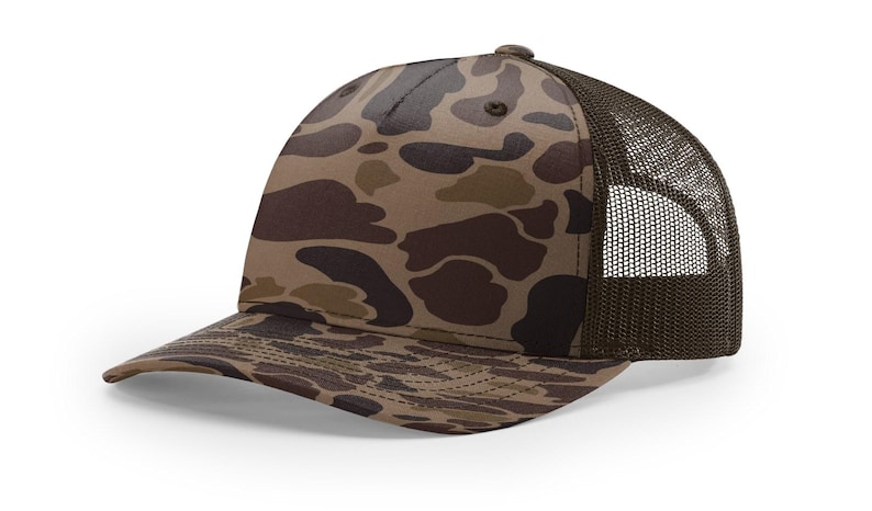 May include: A brown and tan camouflage trucker hat with a curved brim. The front panels feature a duck camouflage pattern, while the back panels are a dark brown mesh. The hat has a standard six-panel design and a button on top.