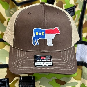 Może przedstawiać: Brązowa i beżowa czapka trucker z haftowaną białą krową z przodu. Krowa trzyma flagę Karoliny Północnej. Czapka ma metkę Richardson 112 z tyłu.