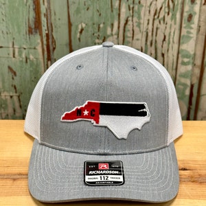 May include: Gray and white trucker hat with a North Carolina state outline patch in red, black, and white. The patch has the letters "N*C" in red. The hat has a black label with the text "EST. 1970 RICHARDSON. ORIGINAL 112 TRUCKER ADJUSTABLE".