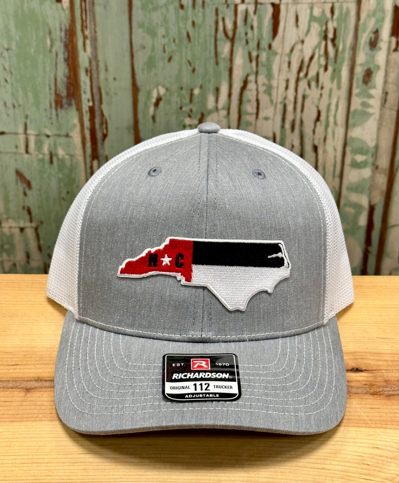 May include: Gray and white trucker hat with a North Carolina state outline patch in red, black, and white. The patch has the letters "N*C" in red. The hat has a black label with the text "EST. 1970 RICHARDSON. ORIGINAL 112 TRUCKER ADJUSTABLE".
