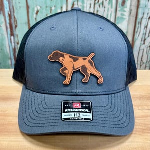 May include: Gray trucker hat with a black mesh back and a brown leather patch featuring a dog silhouette. The hat has a curved brim and an adjustable strap. The Richardson brand label is visible on the front.