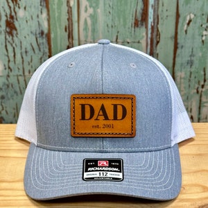 May include: A gray and white trucker hat with a brown leather patch that says "DAD est. 2001". The hat is adjustable and has a Richardson 112 logo.