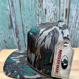 K&ouml;nnte beinhalten: Eine Camouflage-Muster-Cap mit flachem Schirm und braunem Seildetail am Rand. Die Cap hat ein Etikett mit dem "Mossy Oak"-Logo. Das Muster der Cap umfasst Gr&uuml;n-, Braun- und Beiget&ouml;ne.