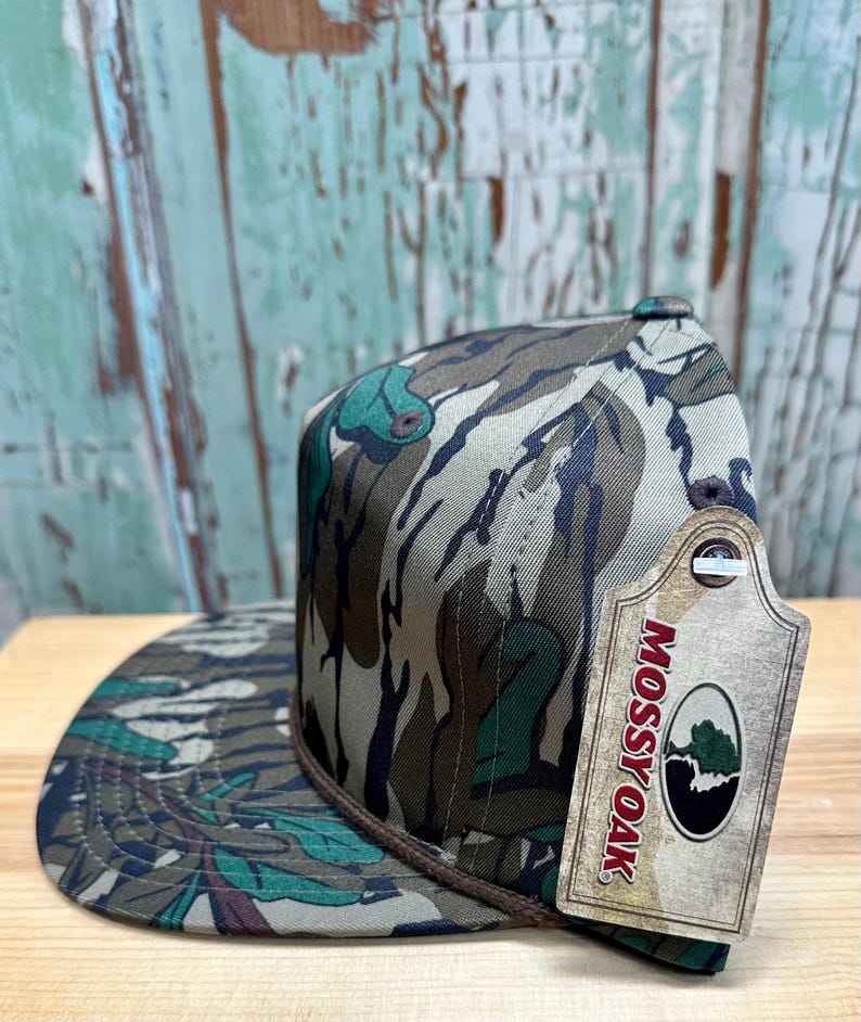 K&ouml;nnte beinhalten: Eine Camouflage-Muster-Cap mit flachem Schirm und braunem Seildetail am Rand. Die Cap hat ein Etikett mit dem "Mossy Oak"-Logo. Das Muster der Cap umfasst Gr&uuml;n-, Braun- und Beiget&ouml;ne.