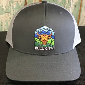 May include: A gray and white trucker hat with a gray bill. The hat features a colorful stained glass style bull head with the text "BULL CITY" underneath.