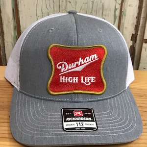 May include: Gray and white trucker hat with a red and gold patch that says "Durham High Life". The hat has a Richardson 112 label on the side.