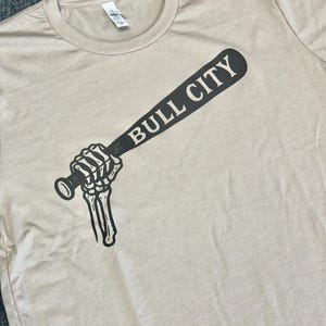 May include: Beige t-shirt featuring a black graphic of a skeleton hand gripping a baseball bat. The bat displays the words "BULL CITY" in a bold, sans-serif font. The shirt has a crew neck and is made of a soft material.
