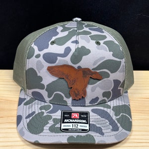 May include: A camouflage trucker hat with a brown leather duck patch. The hat is adjustable and has the text "Richardson" and "112" on the label.