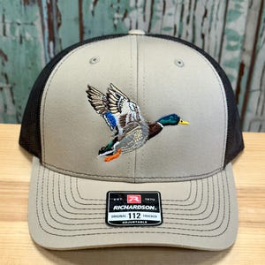 May include: A beige and black trucker hat with an embroidered image of a mallard duck in flight. The hat has a black mesh back and a brown adjustable strap with the text "EST. 1970 RICHARDSON. ORIGINAL 112 TRUCKER ADJUSTABLE".