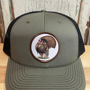 May include: A green and black trucker hat with a brown embroidered patch of a wild turkey. The patch is a circle with a white background.