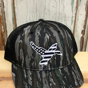 May include: A black and green camouflage trucker hat with a white embroidered duck flying with an American flag design.