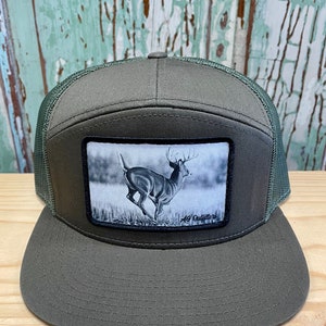 May include: A green and black trucker hat with a patch of a whitetail deer running through a field. The patch has the text "AG Outfitters" on the bottom right corner.